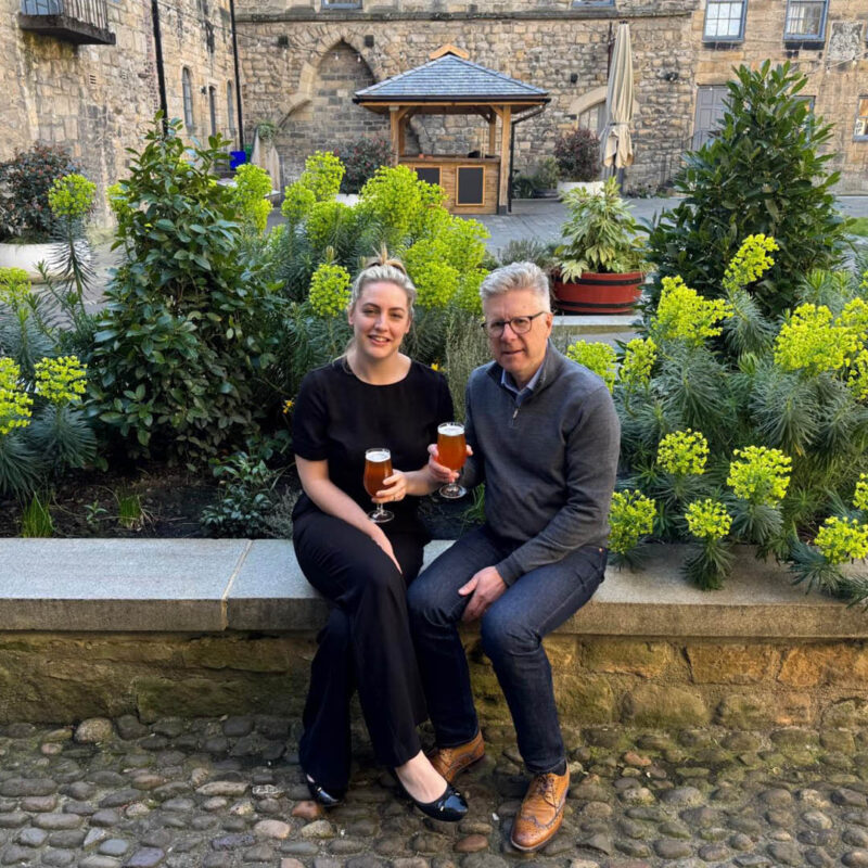 A white woman with blonde hair and an older white man with grey hair sit side by side on a stone wall. Behind them is the beautifully landscaped Cloister Garden of Blackfriars Restaurant. The duo smile at the camera as they each hold up a schooner of beer.