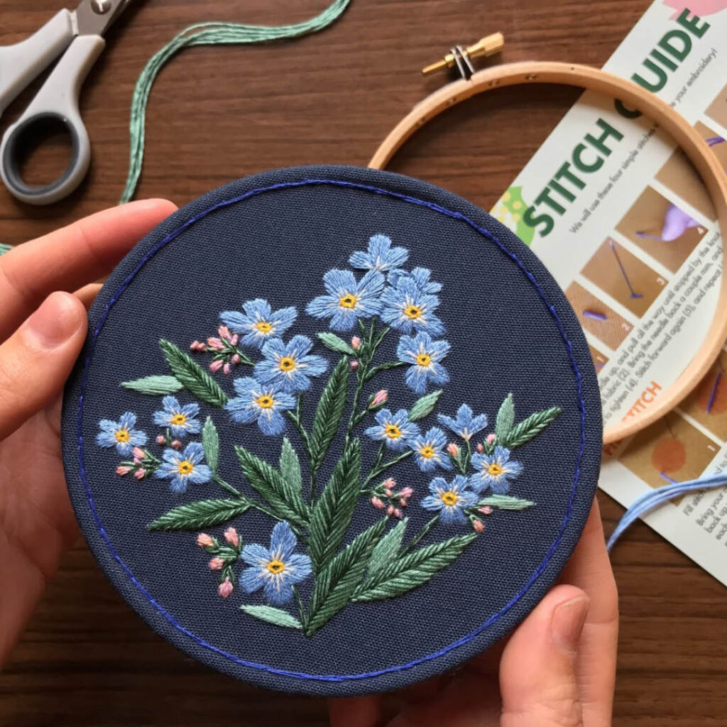 Hands hold a circular piece of embroidery featuring many small blue flowers with yellow centres and green stems.