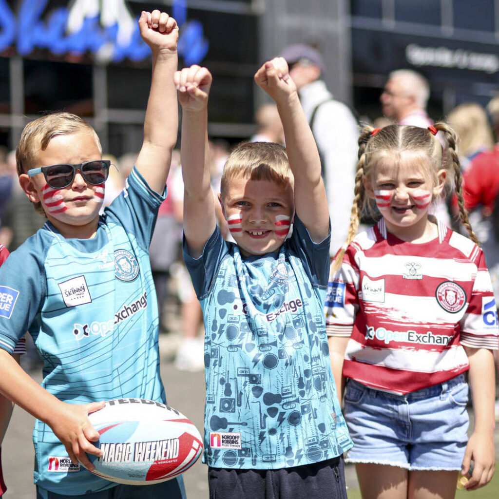 Three little children, two boys and one girl, stand outside of St James' Park. They wear rugby kits and have their faces painted with stripes in the colour of their kits. One boy holds a rugby ball which is printed with the words Magic Weekend. The boys hold their arms up in the air, while the little girl grins at the camera.
