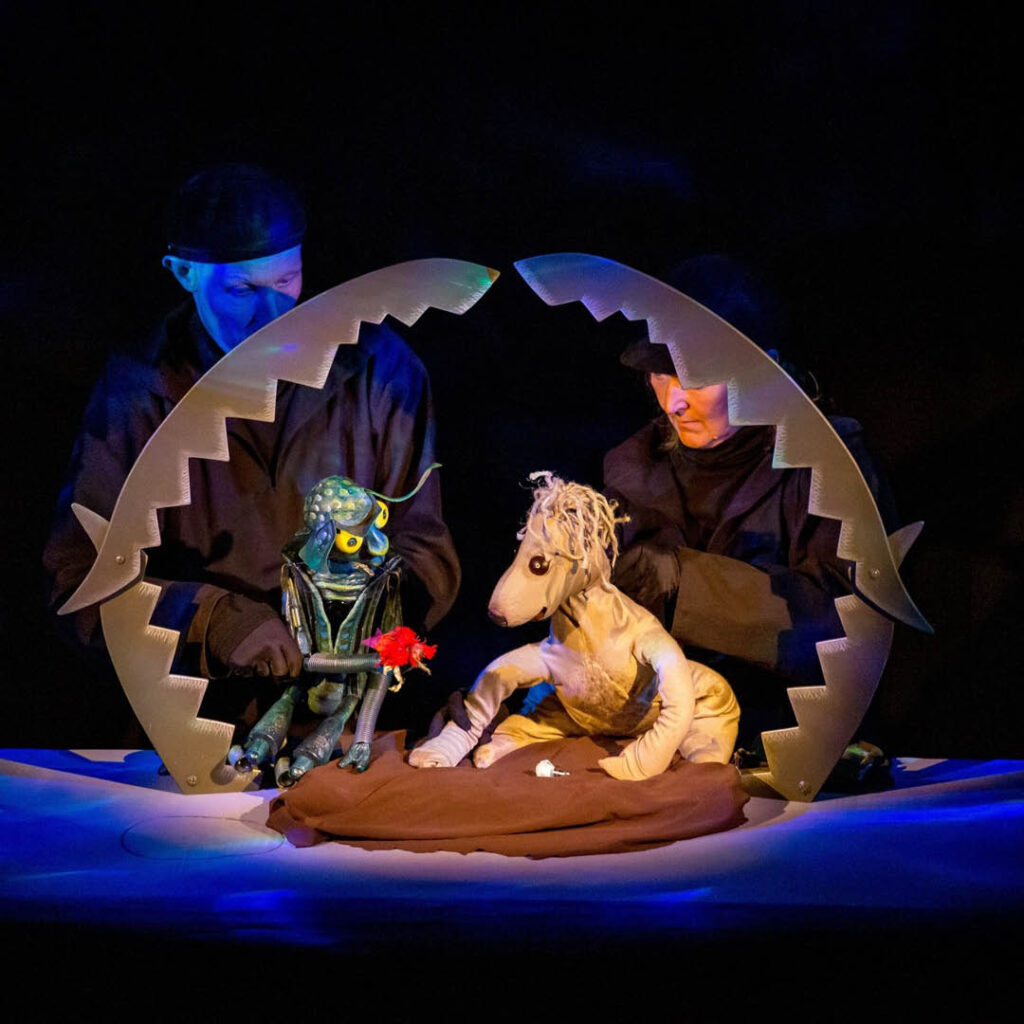 Two strange puppets - one shaped like a robotic bug, the other some kind of lion bear - are positioned on a dark stage. They are perched on a piece of brown fabric above a blue surface, making it look like they are floating on a raft at sea.