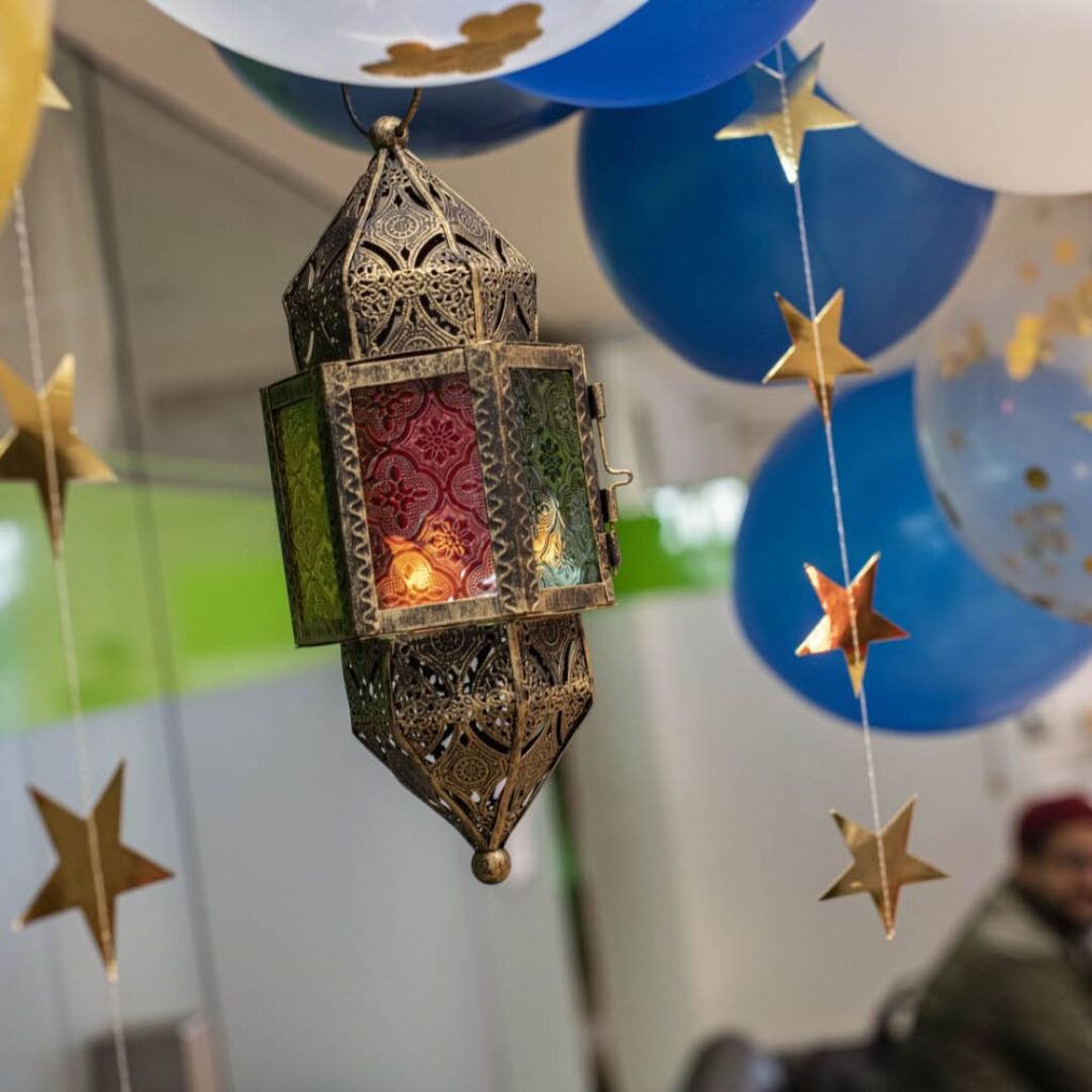 An islamic style lantern hangs from the ceiling. Strings of golden stars and white and blue balloons hang around it. The room has been decorated for the Open Iftar at the BALTIC.