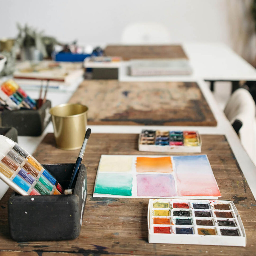 A palette of paints is placed on a wooden board on a table. Next to it is an open sketchbook featuring different coloured painted squares. Various pots and paintbrushes can be seen in the background.