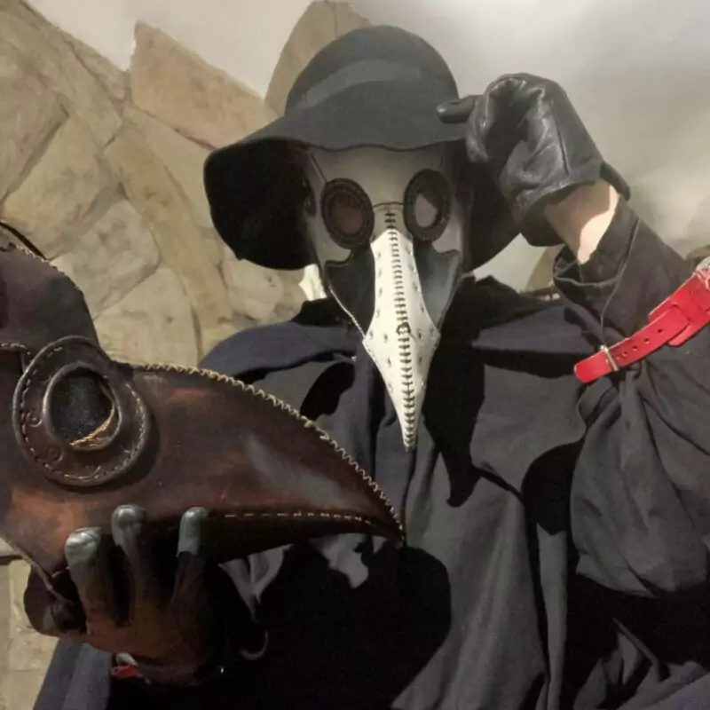 A person dressed as a plague doctor tips their black hat at the camera. In their gloved hand, they hold another brown leather plague mask.