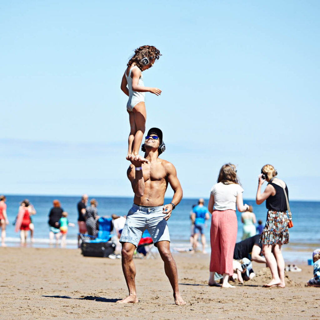 A topless dad on the beach balances his young daughter on his hands. He looks up at her and grins. They both wear headphones. They are enjoying the family session of Silent Disco at the Beach at Tynemouth.