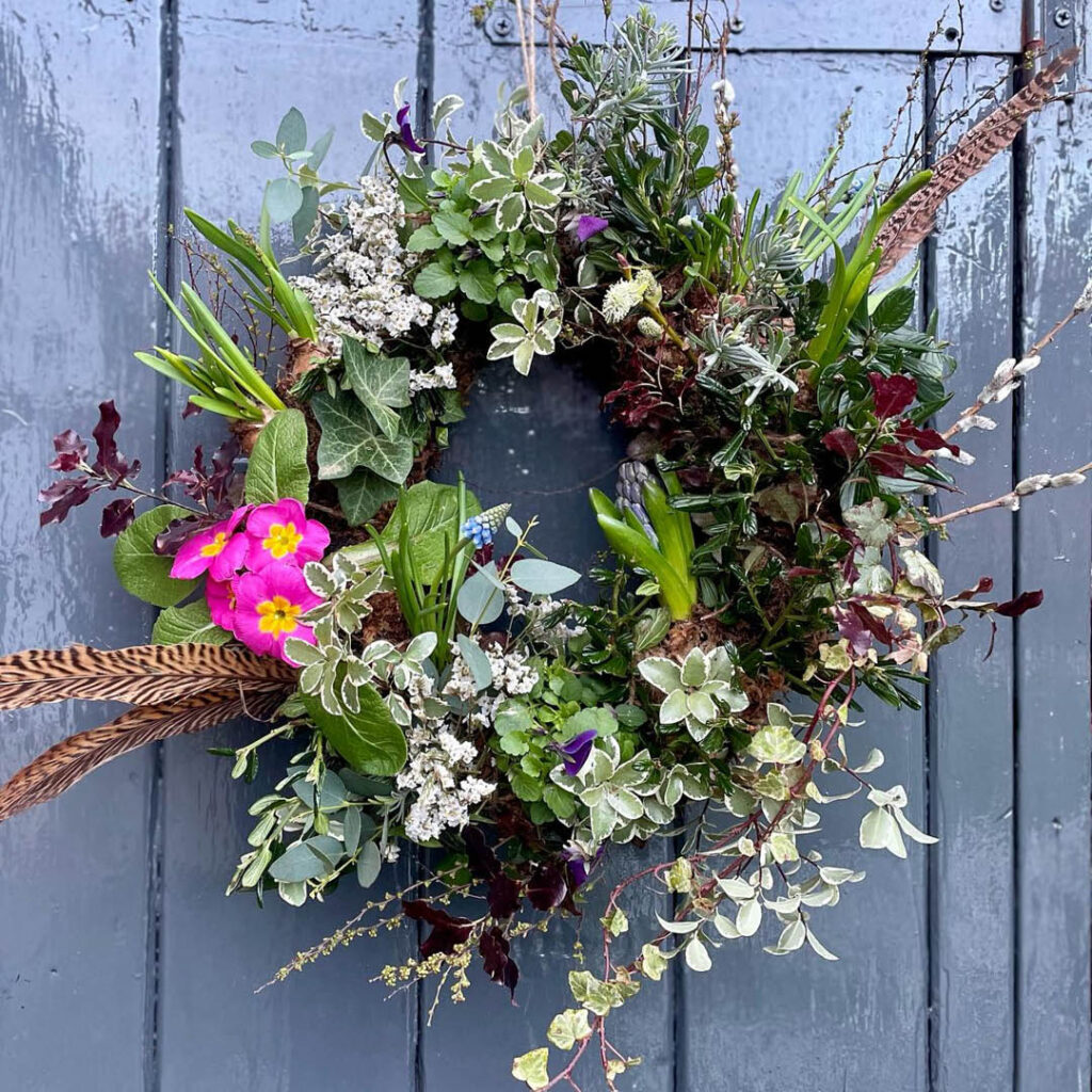 A wreath hangs on a grey wooden door. It is made from various pieces of foliage, flowers and feathers.