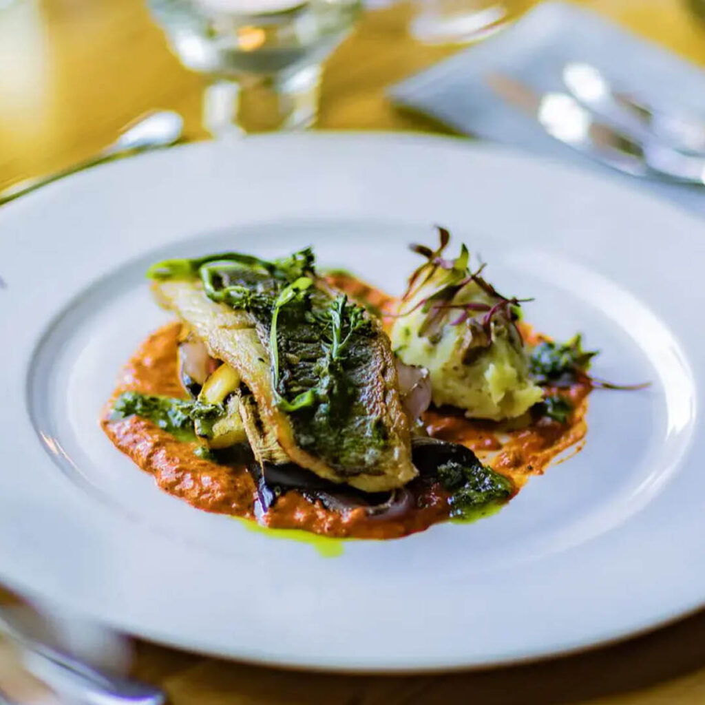 A white plate of roasted fish fillet topped with greens, on a bed of chorizo stew. It is one of the dishes on The Biscuit Factory's Spring Supper Club menu.
