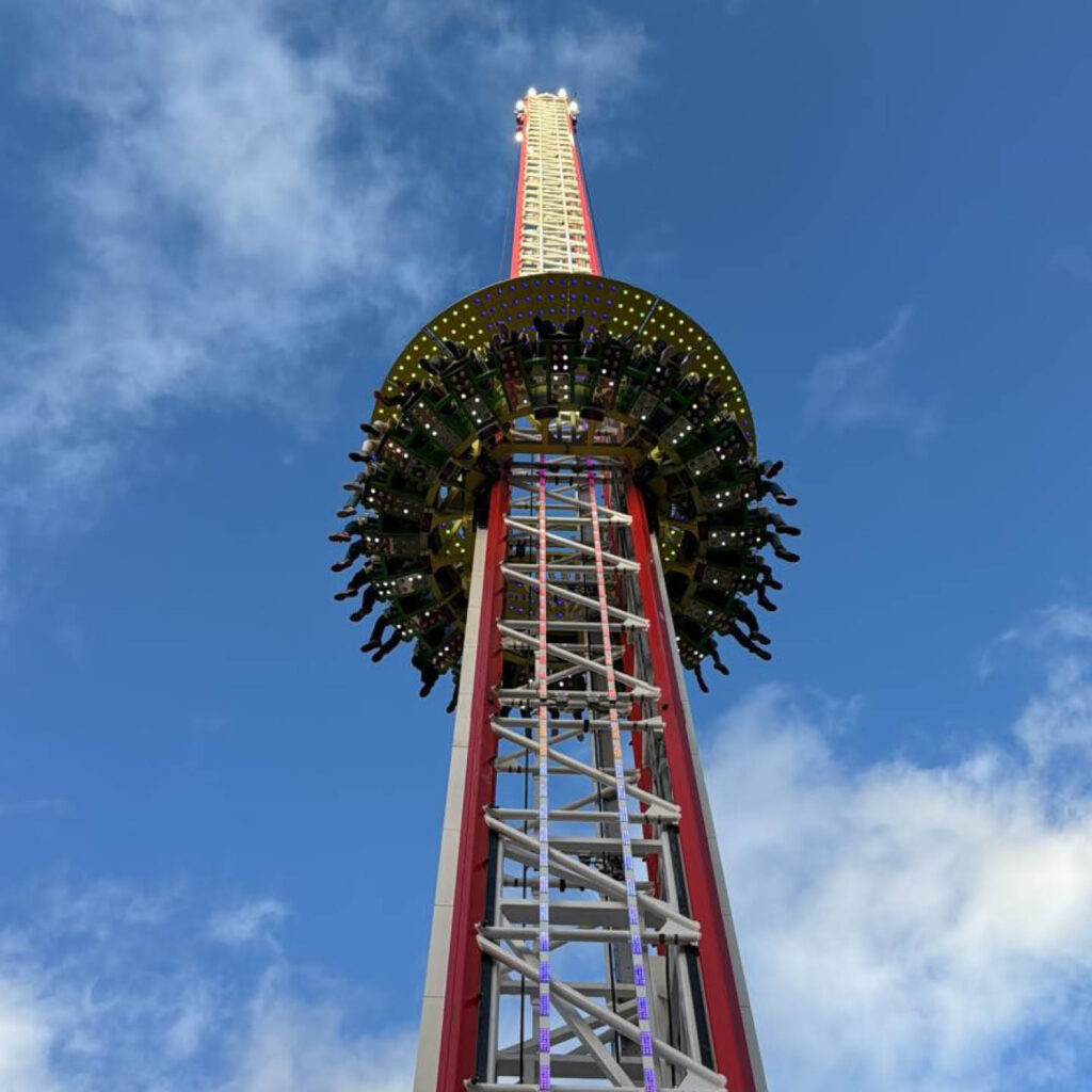 The Drop Tower ride stands against a bright blue sky. People's legs dangle from the seats encircling the middle tower. This is one of the rides at The Hopping funfair this summer.
