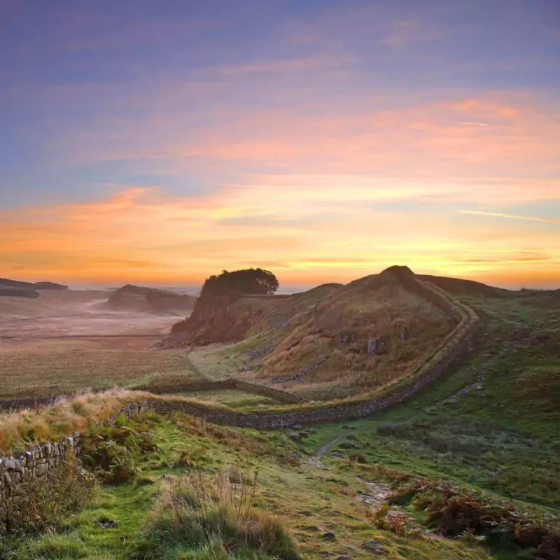Hadrian's Wall curves into the distance between rolling hills and fields. The sky above it is streaked blue, orange and pink with the setting sun.