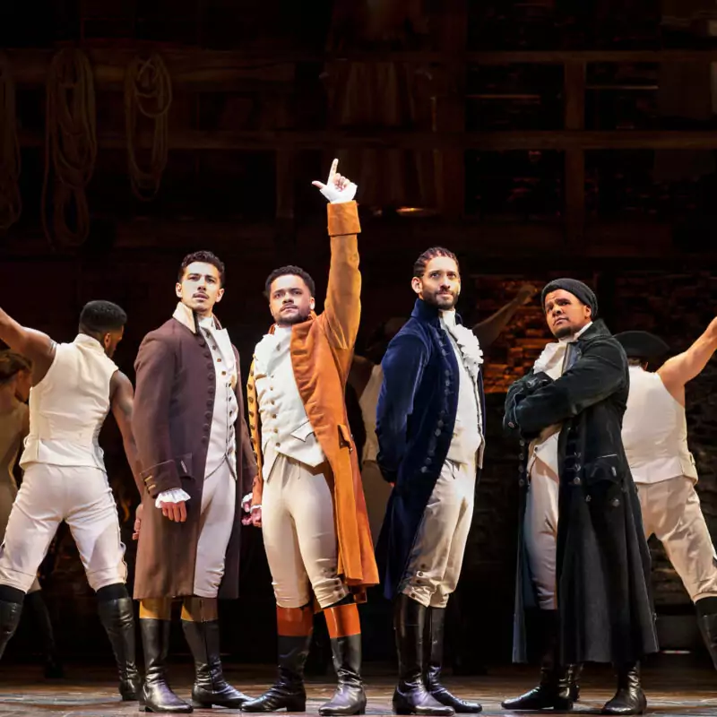 The cast of Hamilton stand on a dark stage. A man wearing an orange trench coat over a white waistcoat and tights points his finger to the sky.