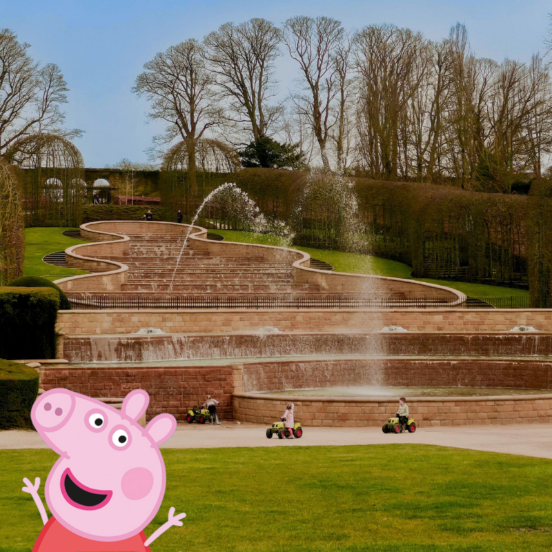 Children ride toy trucks at the bottom of the huge cascading water feature at The Alnwick Garden. Peppa Pig pops out from the bottom left corner, arms raised and a smile on her face.
