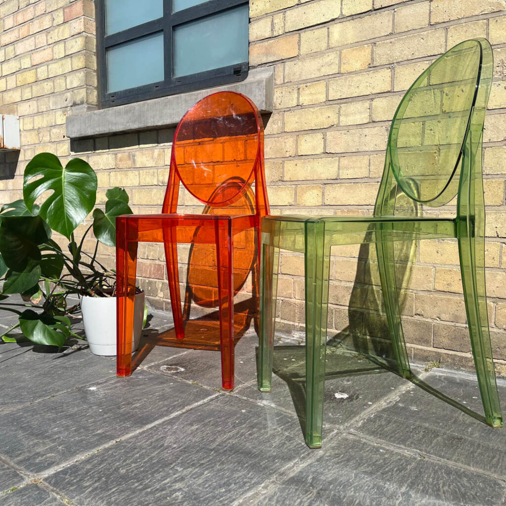 Two designer chairs in clear orange and green plastic are positioned against a brick wall next to a big monstera plant. These chairs are on sale at BALTIC's yard sale.