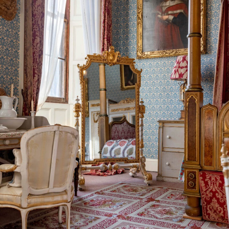 A gold framed mirror stands in the corner of an ornate bedroom in Raby Castle. The wooden posts of a four poster bed can be seen in one corner. A large gold framed portrait hangs from the wall.