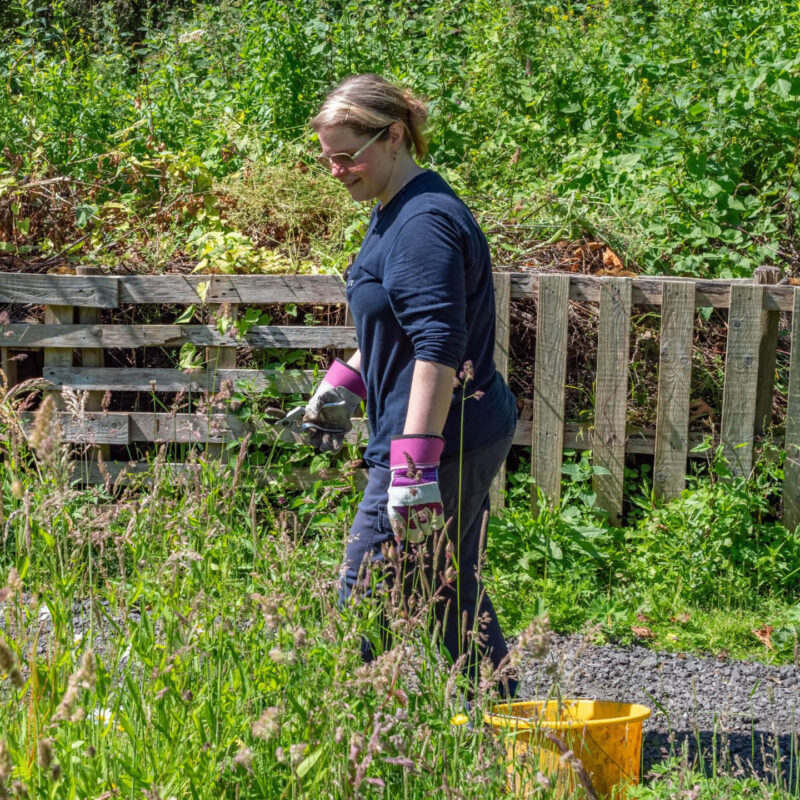 A woman wearing a blue fleece and pink gardening gloves smiles as she walks around a sunny wildlife garden at Washington Wetland Centre.