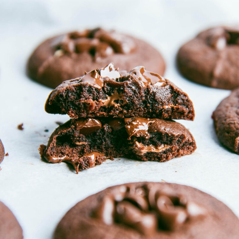 Gooey chocolate chip cookies are lined on a white baking sheet. One of them has been broken in half to reveal the gooey ganache centre.