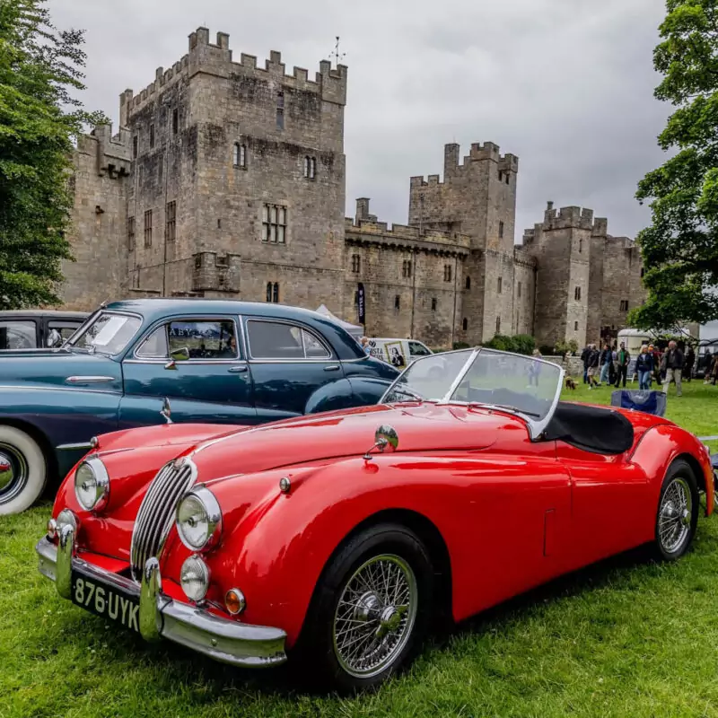 An old red car is parked in the grounds of Raby Castle, which looms behind it. Crowds of visitors can be seen in the distance.