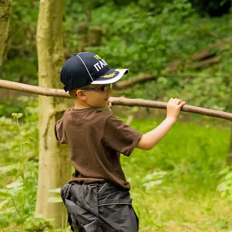 A little boy in a t shirt and cap holds a long stick in both hands. He is building a den at Washington Wetland Centre.