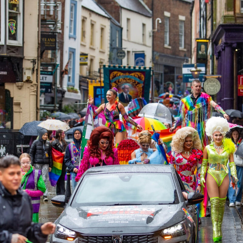 Durham Pride's annual parade. A crowd of people walk down a street in Durham wearing colourful clothes. The crowd is led by four drag queens in vibrant outfits.