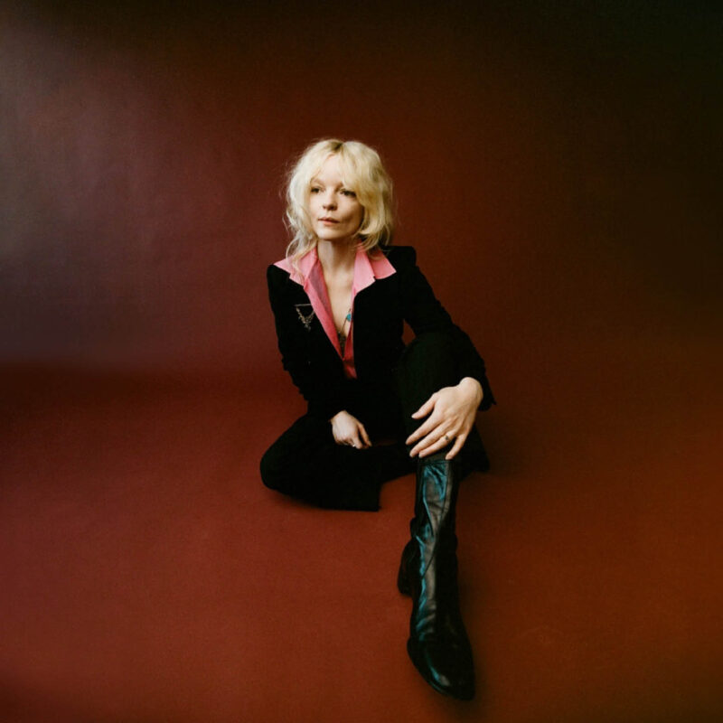 Jessica Pratt is a white woman with bright blonde wavy hair. She wears a black open necked jumpsuit with long black boots. She sits on the floor in a maroon room, looking into the distance.