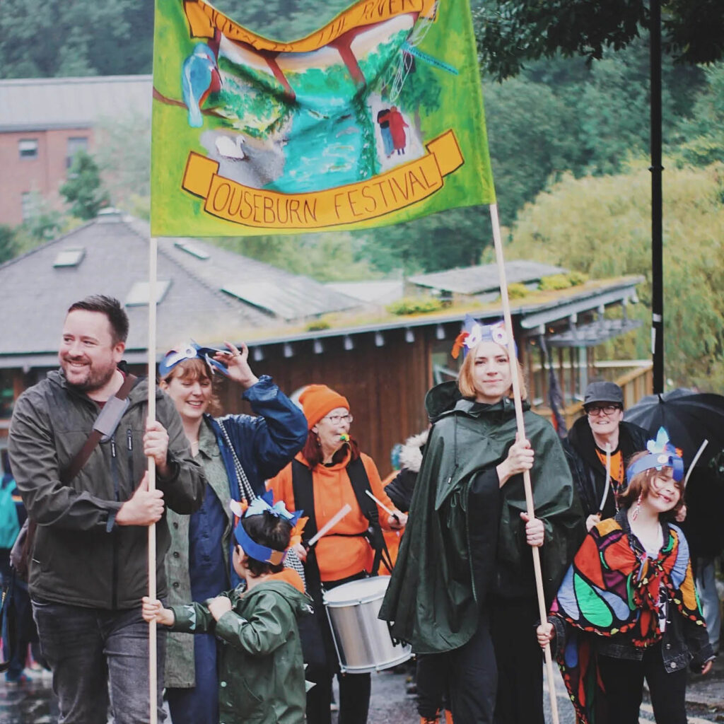 Adults and children wearing raincoats and parade masks grin as they walk along Ouseburn's Lime Street. A pair hold a big green banner between them which says Ouseburn Festival.
