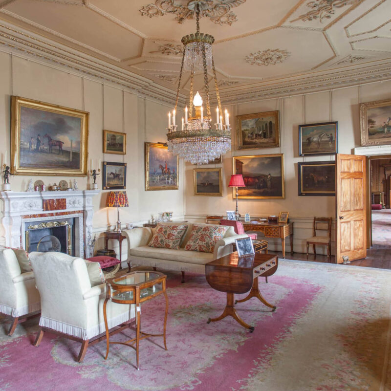 An ornate drawing room in Raby Castle. A crystal chandelier hangs at the centre of the room above a white sofa and chairs. Landscape paintings adorn the walls.