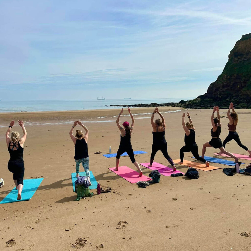 Seven woman raise their arms above their heads in a yoga pose. They stand on colourful yoga mats laid out on golden sand at St Edward's Bay. The sea and sky is blue around them.
