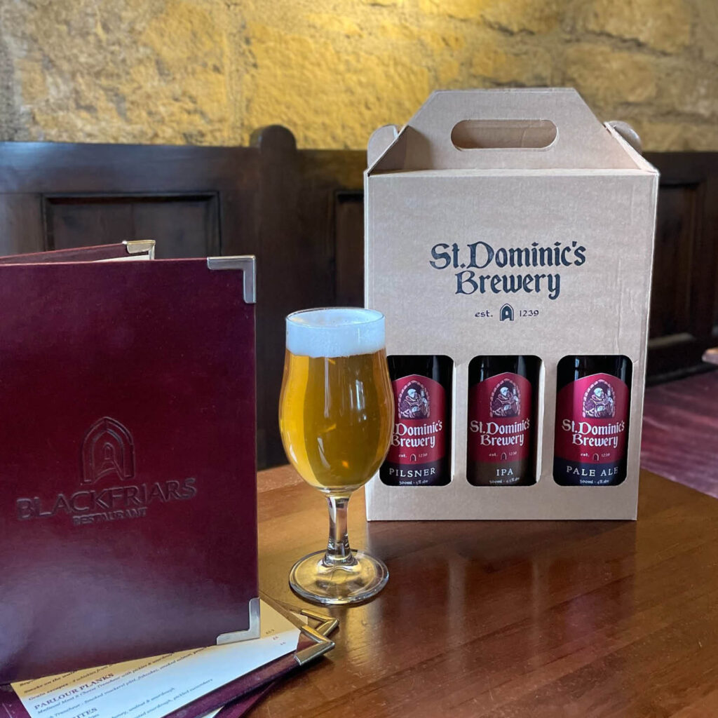 A stemmed glass of beer next to a cardboard box of three beer bottles with the St Dominics Brewery branding. A red Blackfriar's Restaurant menu stands next to them.