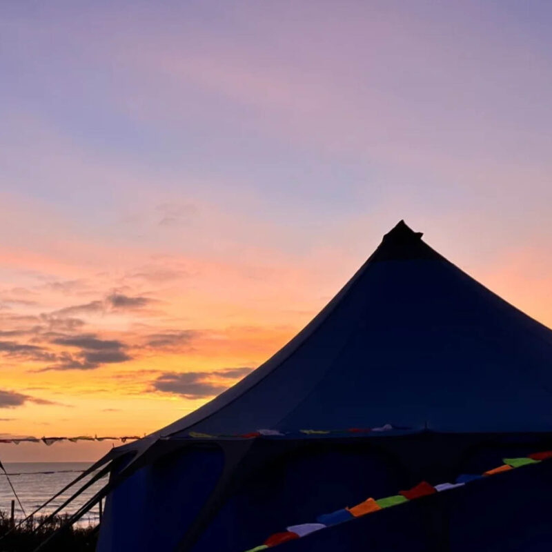 A large tent with bunting is silhouetted against a purple and orange sunset sky. It is one of the tents at the North Sea Wellbeing Weekender.