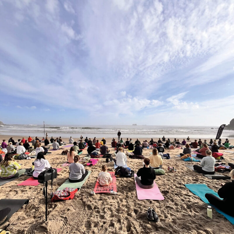 People sit and stretch on yoga mats laid out on a beach beneath a blue sky.