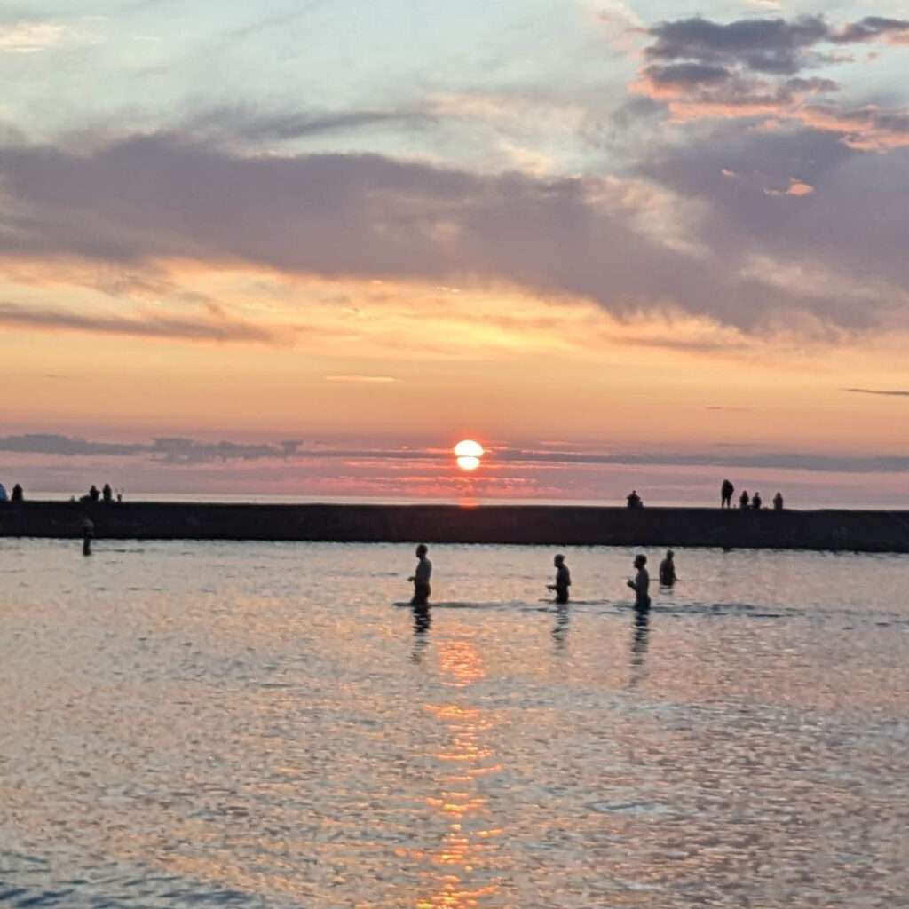 Silhouettes of people wading into the calm sea at Cullercoats Beach. The sun emerges from the horizon and streaks the sky orange.
