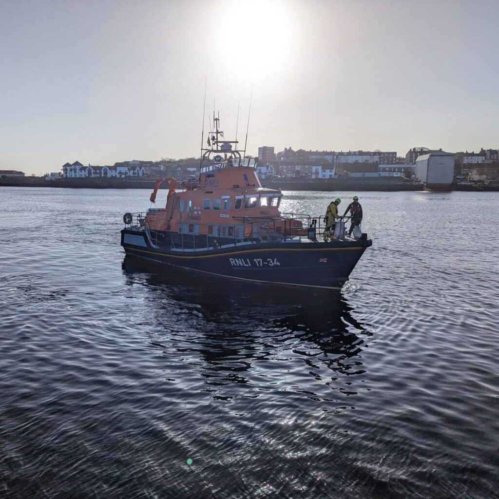 An RNLI lifeboat floats in the sea. Two volunteers stand on its deck. Tynemouth waterfront can be seen in the distance beneath a shining white sun.