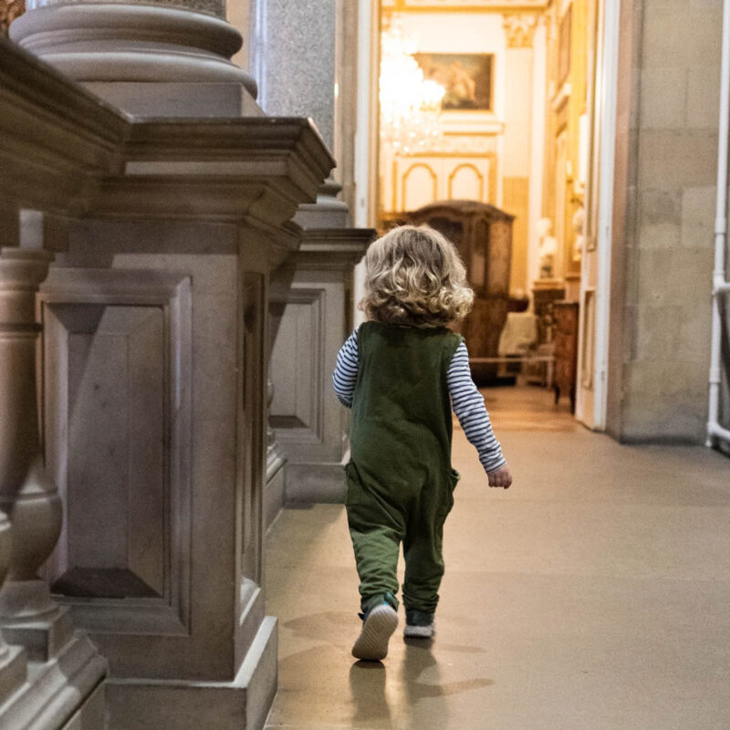 A blond haired toddler in a green one piece runs along a hallway inside The Bowes Museum. They are taking part in the Bug Hunt activity.