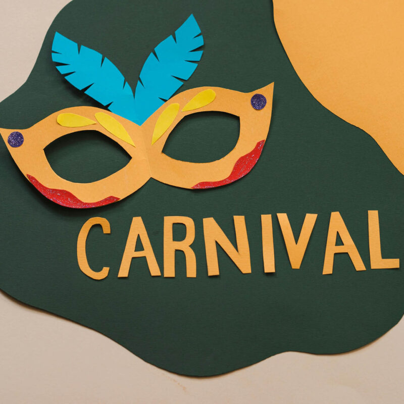 A yellow carnival mask with blue paper feathers. It is made out of paper. Below it, yellow paper has been cut out to spell the word 'Carnival'.