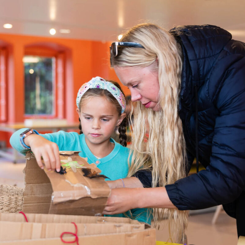 A little girl wearing a turquoise top and a headband sticks something down on a piece of cardboard that her mum holds for her. She is taking part in the Bird Habitats craft workshop at The Bowes Museum.