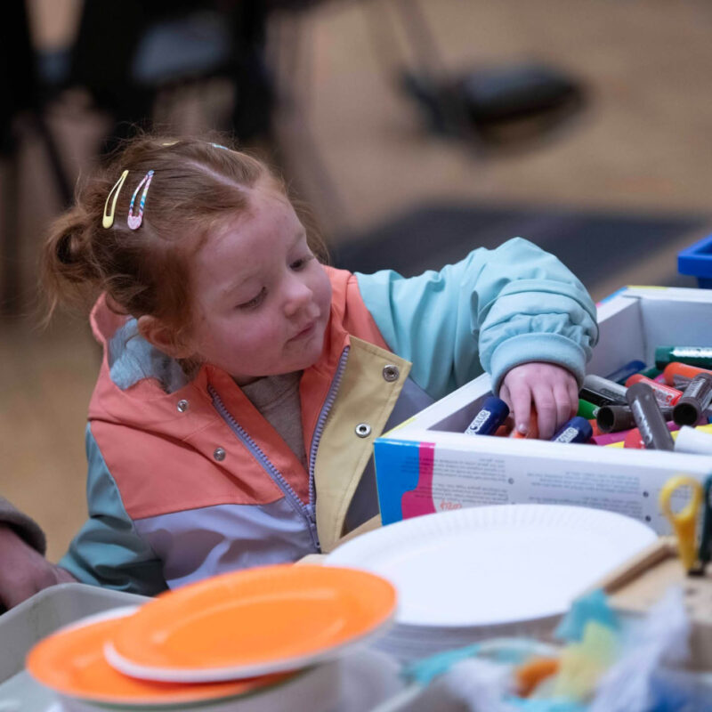 A little girl with bunches and clips in her hair chooses a pen from a big tray. She is taking part in a Crafty Kids session at Shipley Art Gallery.