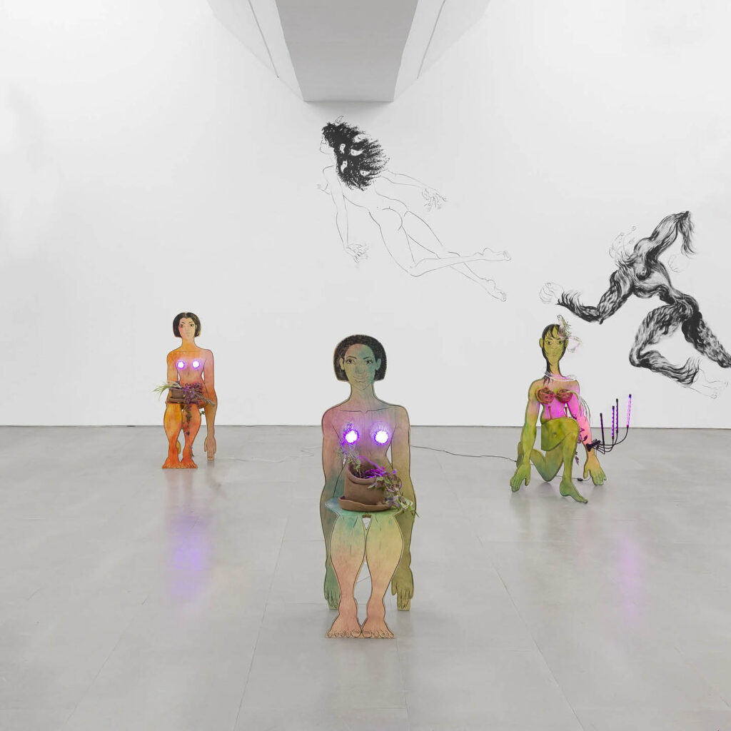 Three sculptures are positioned in a triangle formation in a white gallery space. They depict smiling human forms holding flower pots. On the wall behind them, black line drawings show a hairy looking human form leaping towards a flying female figure. This is part of Saelia Aparicio's exhibition at BALTIC: A Joyful Parasite.